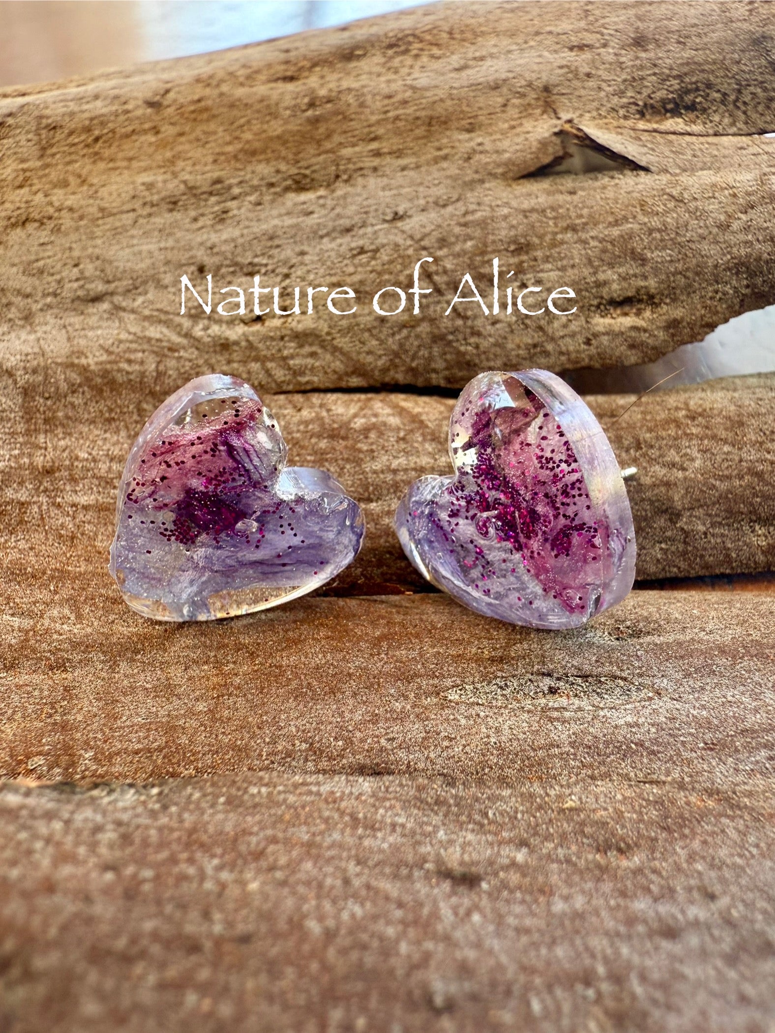 Heart-shaped purple and pink earrings on bark with 'Nature of Alice' branding.